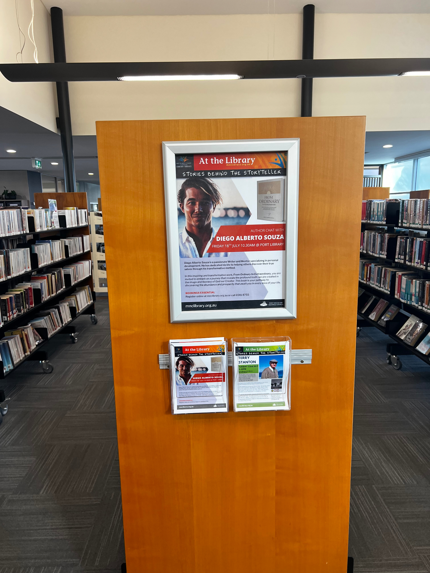 Cartaz do evento exposto no interior da Biblioteca de Port Macquarie, entre as estantes de livros.
